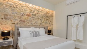 A bedroom with a stone wall, a bed with white bedding, two bedside lamps, and a robe rack with white robes. - THIRAMENOUS RESIDENCE - FIVOS SUITE (Athina)