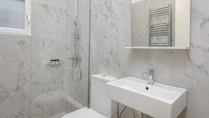 A modern bathroom with a glass-enclosed shower, a white sink, and a marble-tiled wall. - THIRAMENOUS RESIDENCE - FIVOS SUITE (Athina)