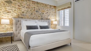 A bedroom with a stone wall, a large bed with white bedding, two bedside tables with lamps, and a window with blinds. - THIRAMENOUS RESIDENCE - FIVOS SUITE (Athina)