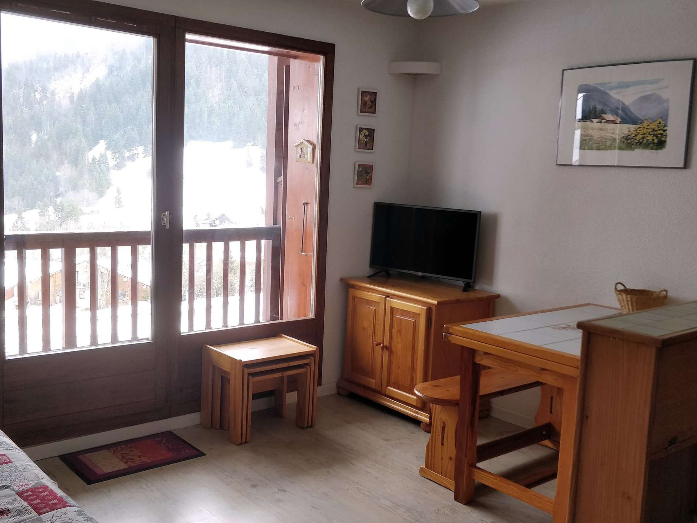 Una habitación con un escritorio de madera, un televisor sobre un mueble bajo, un taburete pequeño y un ventanal con vistas a un paisaje nevado.