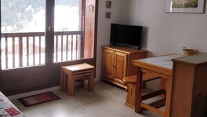 Een kamer met een houten bureau, een televisie op een kastje, een klein krukje en een groot raam met uitzicht op een besneeerd landschap.
