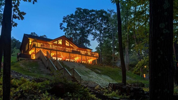 A large, two-story wooden cabin with a balcony, surrounded by a forested area.