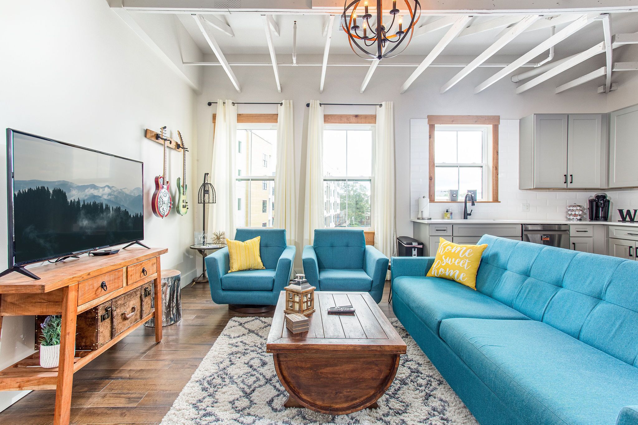 Una sala de estar con televisor, sofás azules, mesa de centro de madera y una cocina al fondo.