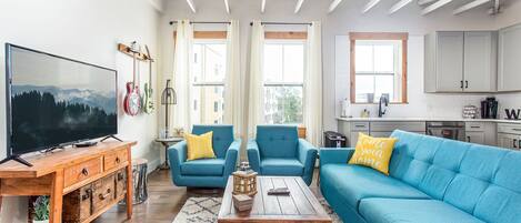 Una sala de estar con televisor, sofás azules, mesa de centro de madera y una cocina al fondo.