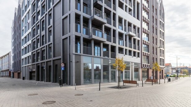 A modern multi-story building with balconies, a paved ground floor, and a clear sky.