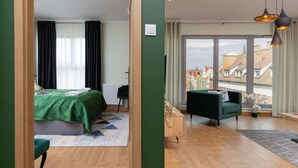 A modern bedroom with a bed, a green chair, a wooden cabinet, and a view of buildings through large windows.