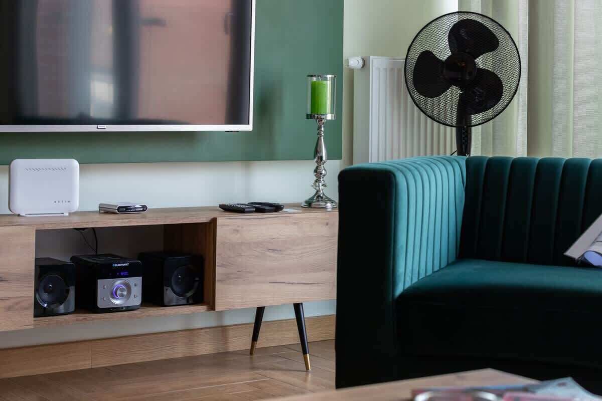 Un salon moderne avec un canapé de couleur turquoise, un meuble de rangement en bois intégrant des haut-parleurs, un téléviseur à écran plat et un ventilateur sur pied.