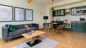 A modern living room with a grey sofa, a wooden coffee table, and a dining area with a round table and chairs. The kitchen features green cabinets and stainless steel appliances.