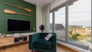A modern living room with a large flat-screen TV, wooden paddleboards on the wall, a green velvet armchair, and a view of a river and buildings through large windows.