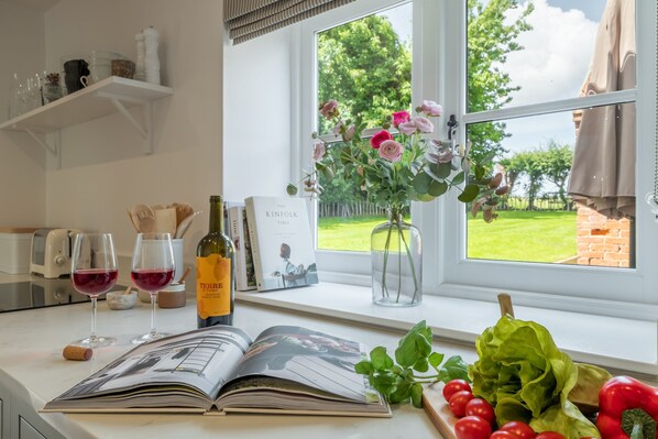 A kitchen counter with a cookbook, wine glasses, a bottle of wine, a vase with flowers, and fresh vegetables.