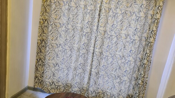 Ein Wohnzimmer mit einem gemusterten Vorhang, einem runden Holztisch mit einer verzierten Platte, einem grauen Sofa mit einem gelben Kissen und einem kleinen runden Tisch mit schwarzem Metallgestell.