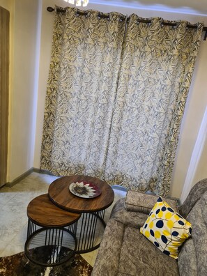 A living room with a patterned curtain, a round wooden table with a decorative plate, a gray sofa with a yellow pillow, and a small round table with a black metal frame. - Homely and safe environment (Nairobi)