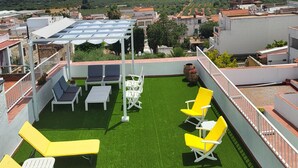 Een terras op het dak met groen kunstgras, gele en witte lounge stoelen en een pergola voor de schaduw.
