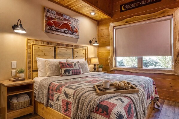 A cozy wooden room with a bed, a nightstand, a lamp, a window with a view, and a sign on the wall.
