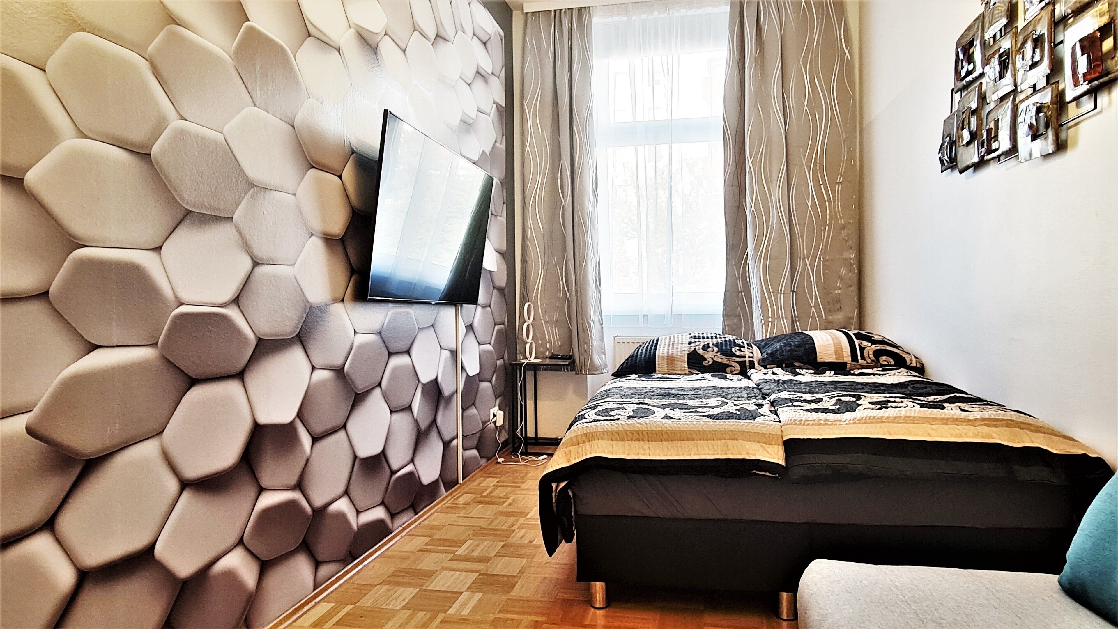 Un dormitori modern amb un disseny de paret hexagonal, un televisió de pantalla plana, un llit amb roba de llit estampada i una finestra amb cortines.