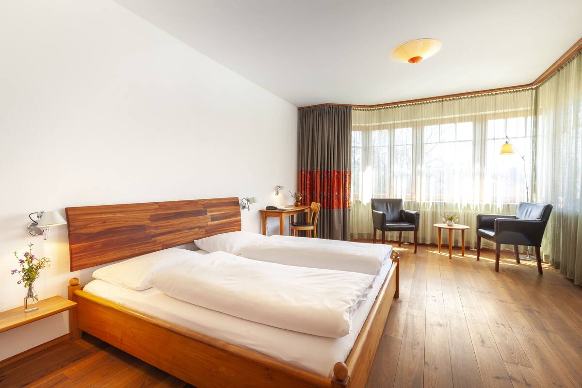 A hotel room with a wooden bed, a desk, two chairs, a small table, and a window with curtains.