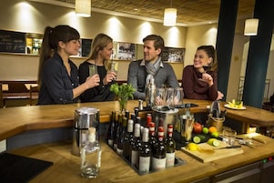 Una zona de bar con mostrador de madera, botellas de vino, copas y una variedad de frutas y verduras sobre la mesa.