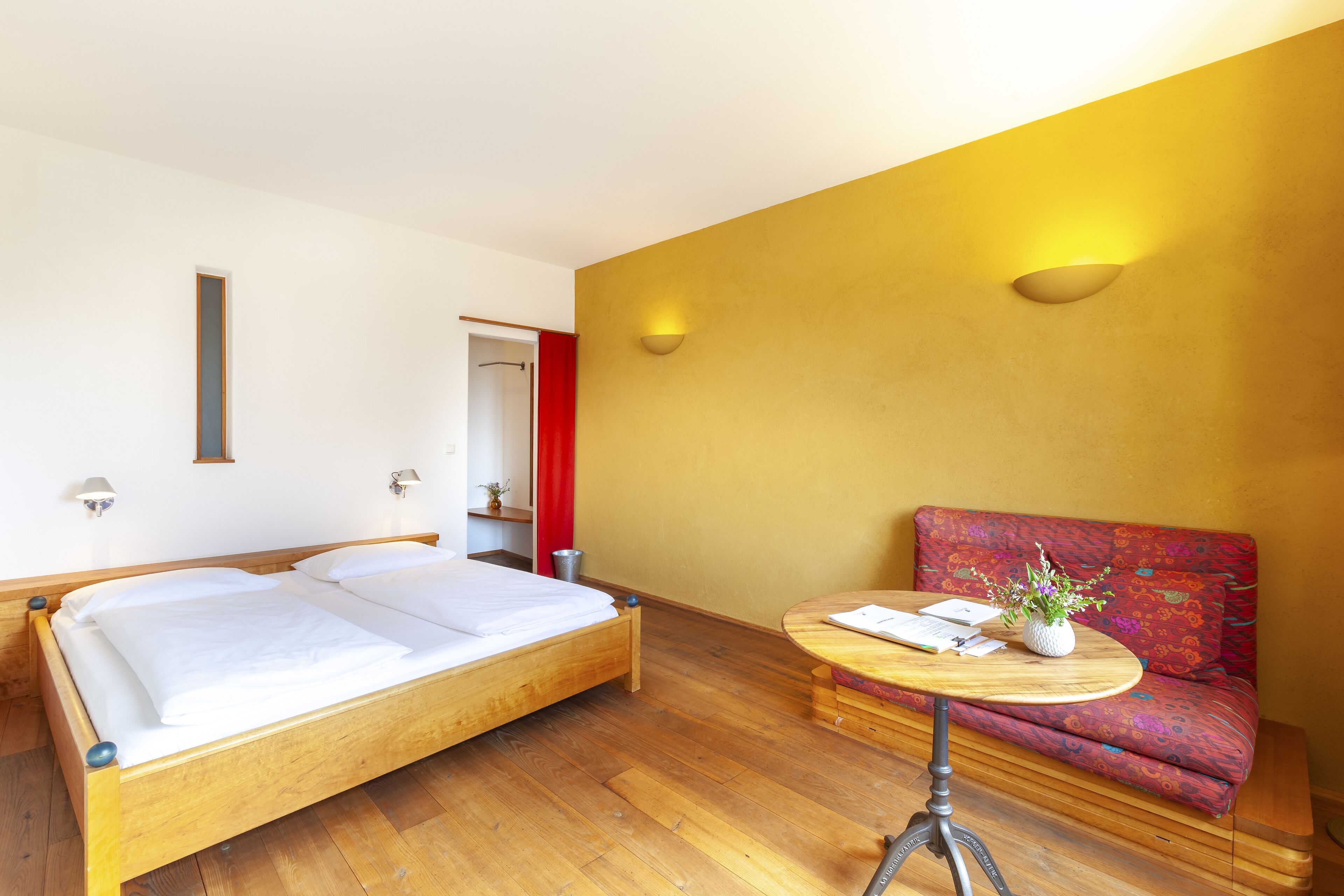 Ein Zimmer mit einem hölzernen Bett, einem kleinen runden Tisch mit einer Vase und einem roten Sofa.