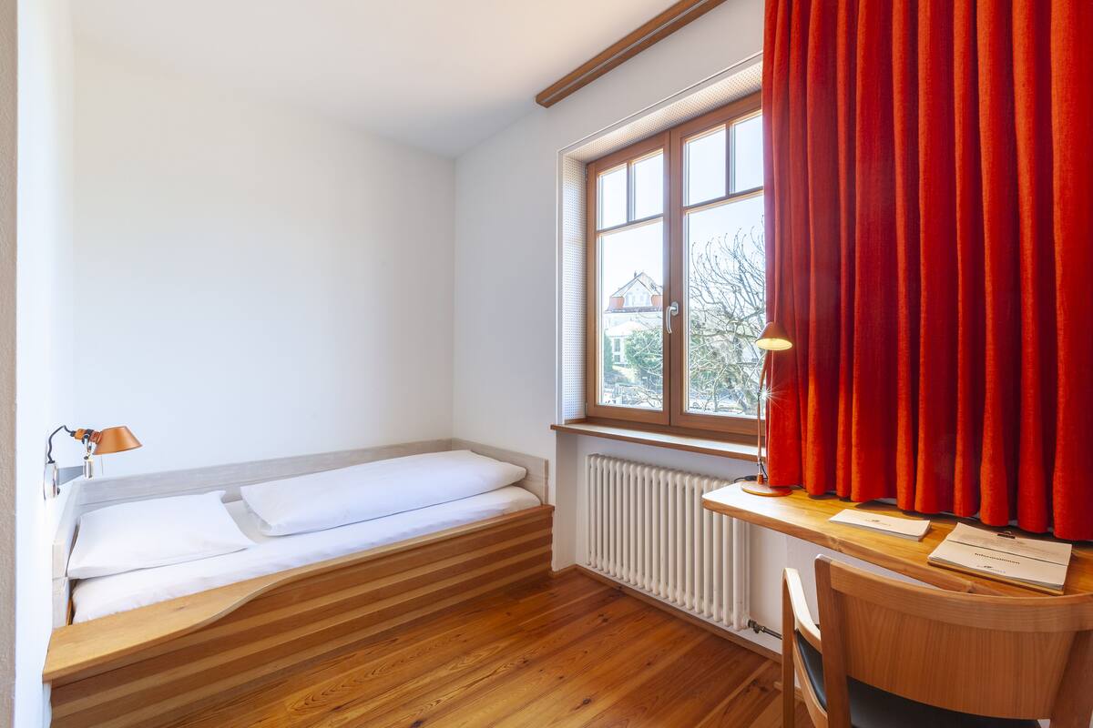 A small, bright room with a wooden bed, a desk with a lamp, and a window with red curtains.