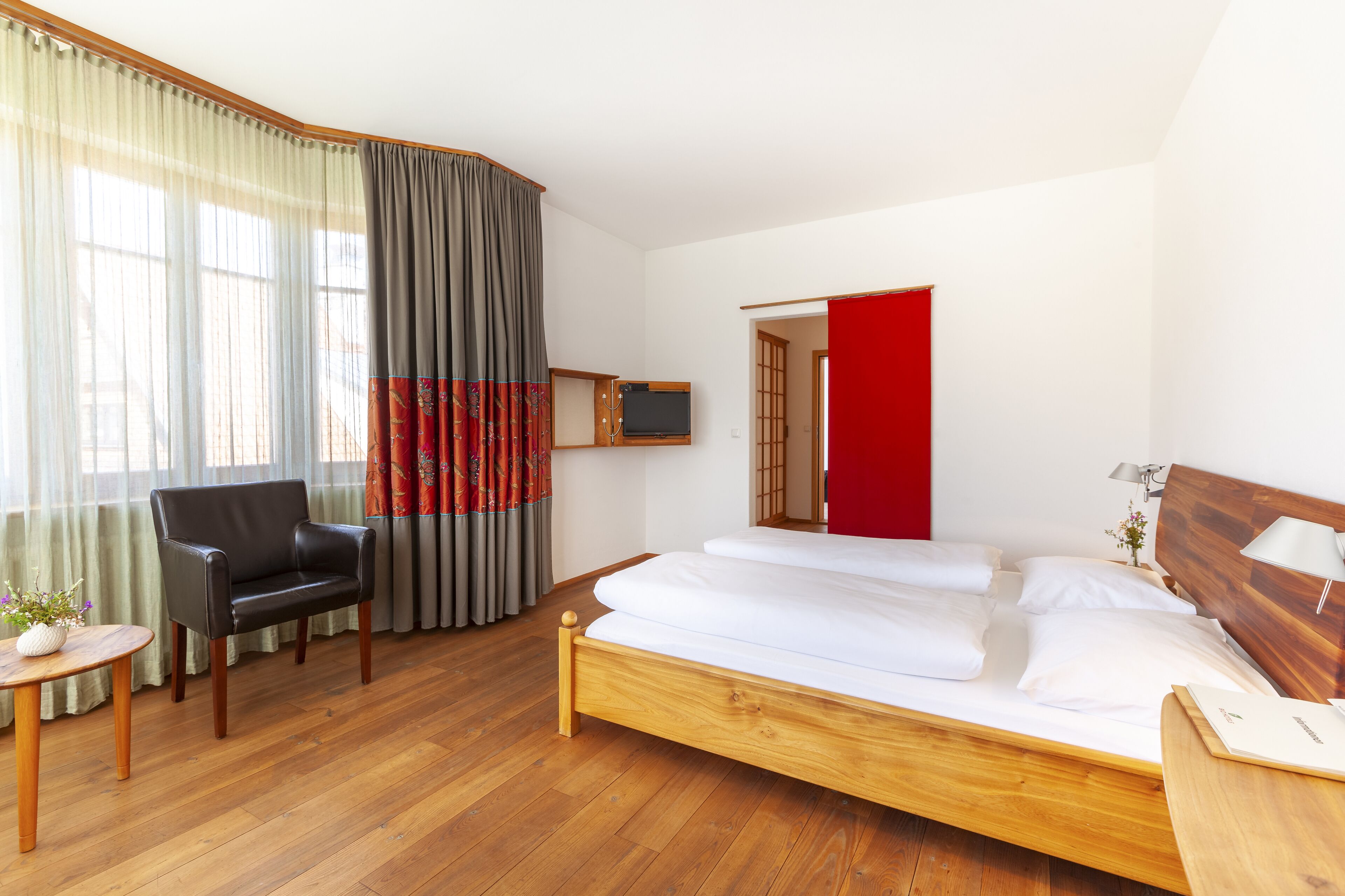A hotel room with a wooden bed, a black leather chair, a small table with a vase, and a TV on a wooden stand.
