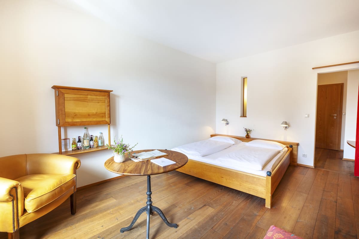 A bedroom with a wooden bed, a leather armchair, a small round table, and a shelf with bottles.