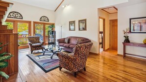 A spacious living room with a wooden floor, a brown leather sofa, patterned armchairs, a glass coffee table, and a bookshelf.