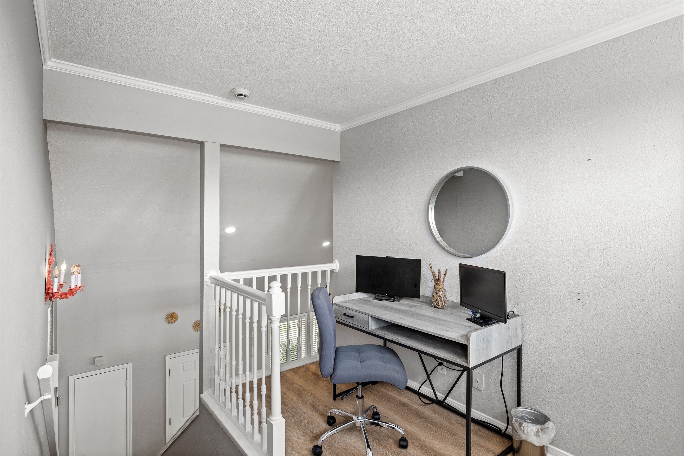 A room with a desk, two monitors, a chair, a round mirror, and a white railing.