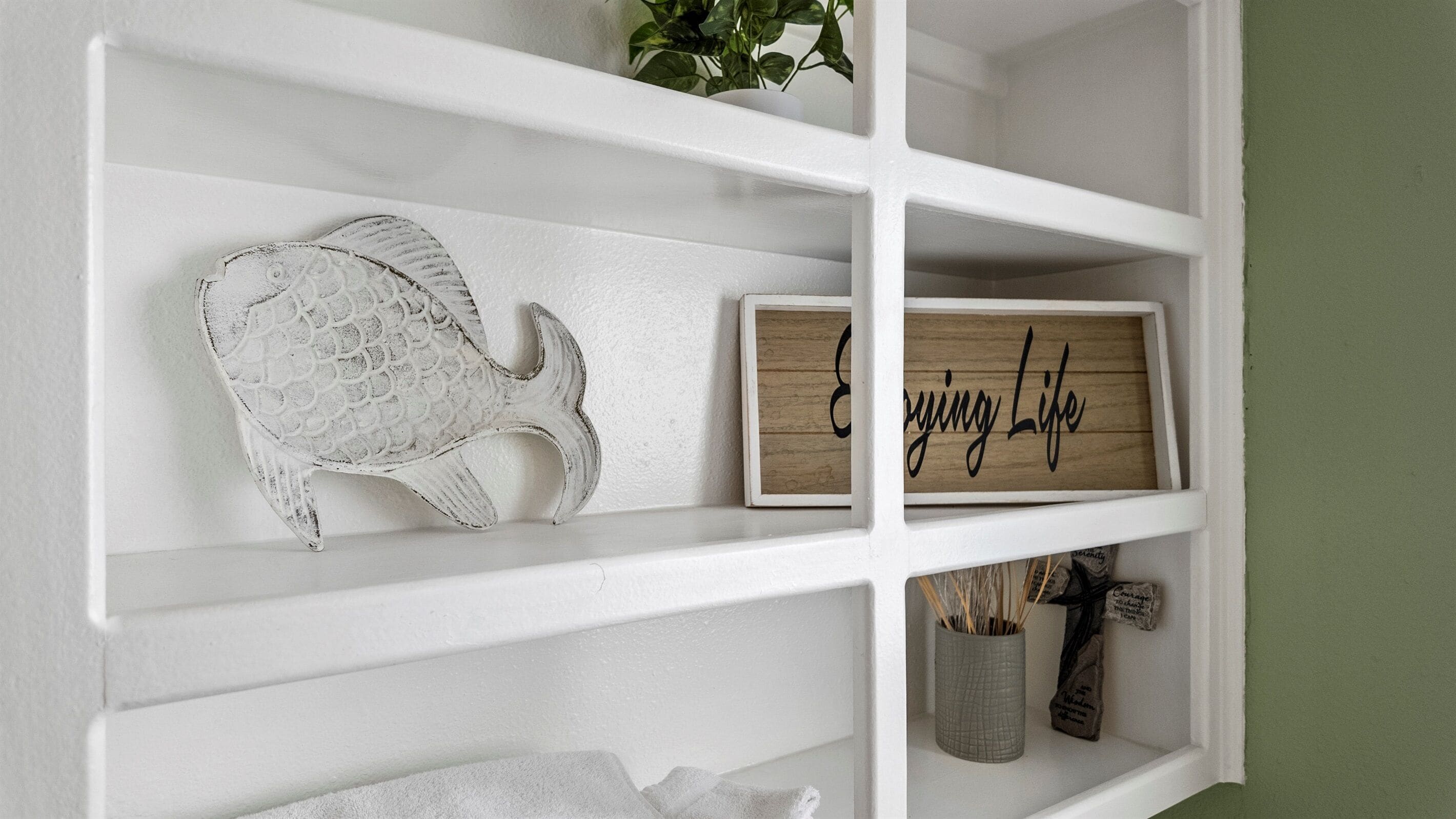 En hvid hylde med en dekorativ fisk, et træskilt med teksten 'Living Life' og en lille vase med pinde.