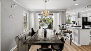 Un comedor con mesa de madera, sillas y una araña. Hay una cocina con refrigerador y televisor.