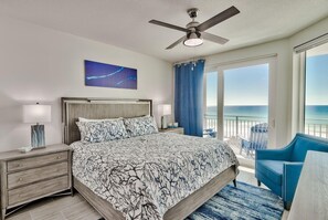 A bedroom with a bed, a ceiling fan, a nightstand, a lamp, a window with a view of the ocean, and a blue chair.