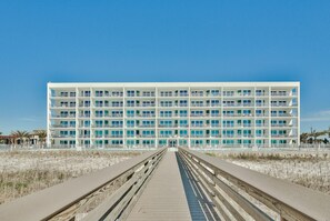 A multi-story hotel with a central entrance, a wooden boardwalk leading to it, and a beach in the background.