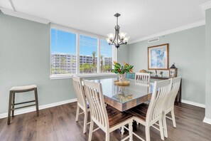 Un coin repas avec une table à plateau de verre, des chaises blanches, un lustre, une fenêtre avec des stores et un tableau encadré au mur.