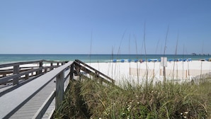 Ein hölzerner Steg führt zu einem Sandstrand mit blauen Sonnenschirmen.