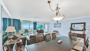 A spacious living room with a large wooden dining table, comfortable seating, and a view of the ocean through large windows.