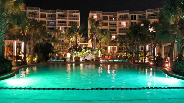 A nighttime view of a hotel pool area with a clear pool, palm trees, and a building with multiple balconies in the background. - Heron 604-Watch for Dolphins from your balcony!  3 BR Condo with Great Views (Fort Walton Beach)