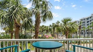 A balcony with a round table and two chairs, surrounded by palm trees and a view of a pool and buildings.