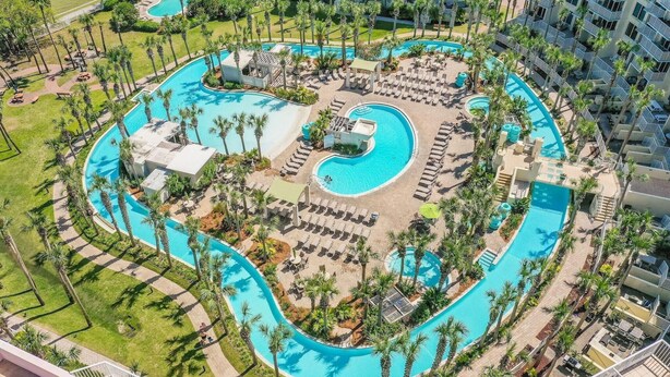 A large, winding pool surrounded by palm trees and sun loungers, with a building complex in the background.
