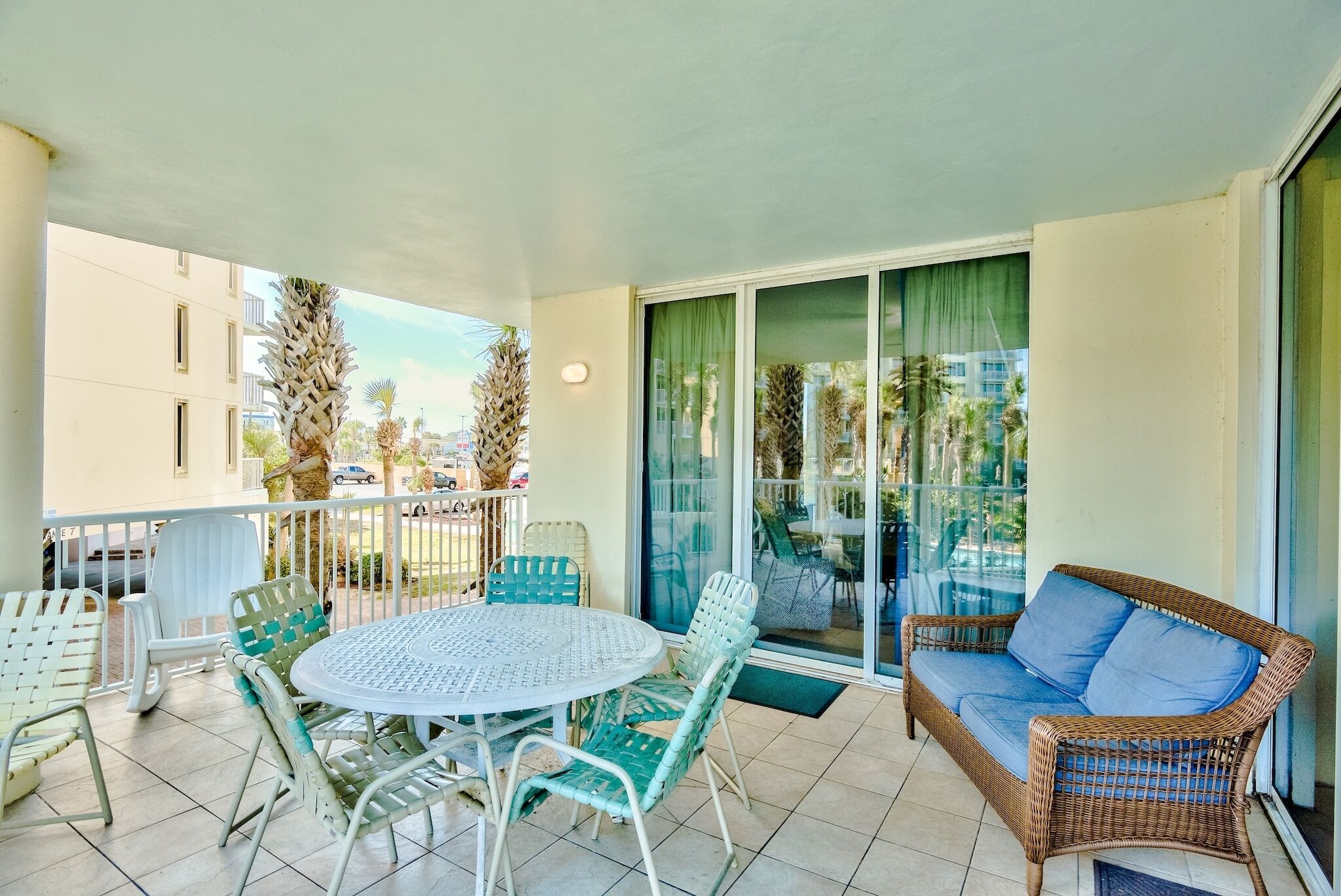 A balcony with a round table and chairs, a wicker sofa, and a sliding glass door leading to a pool area.