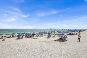En strand med rader av parasoller och strandstolar, en pir i bakgrunden och människor som kopplar av.