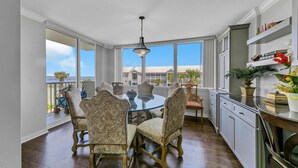 A dining area with a round glass table and four chairs, large windows with a view of buildings, and a kitchen with white cabinets and a countertop.