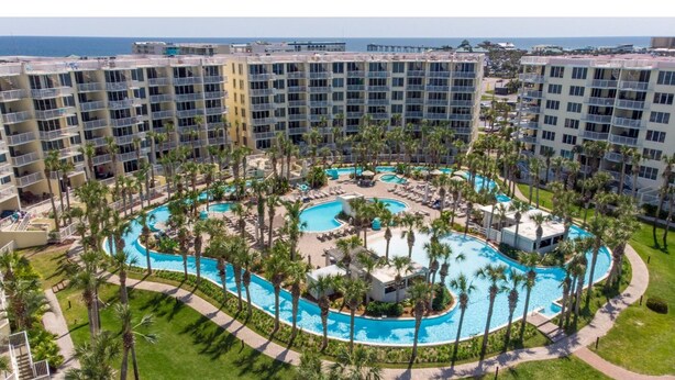 Een hotel met meerdere verdiepingen, een centraal gelegen zwembad omgeven door palmbomen en uitzicht op de oceaan.