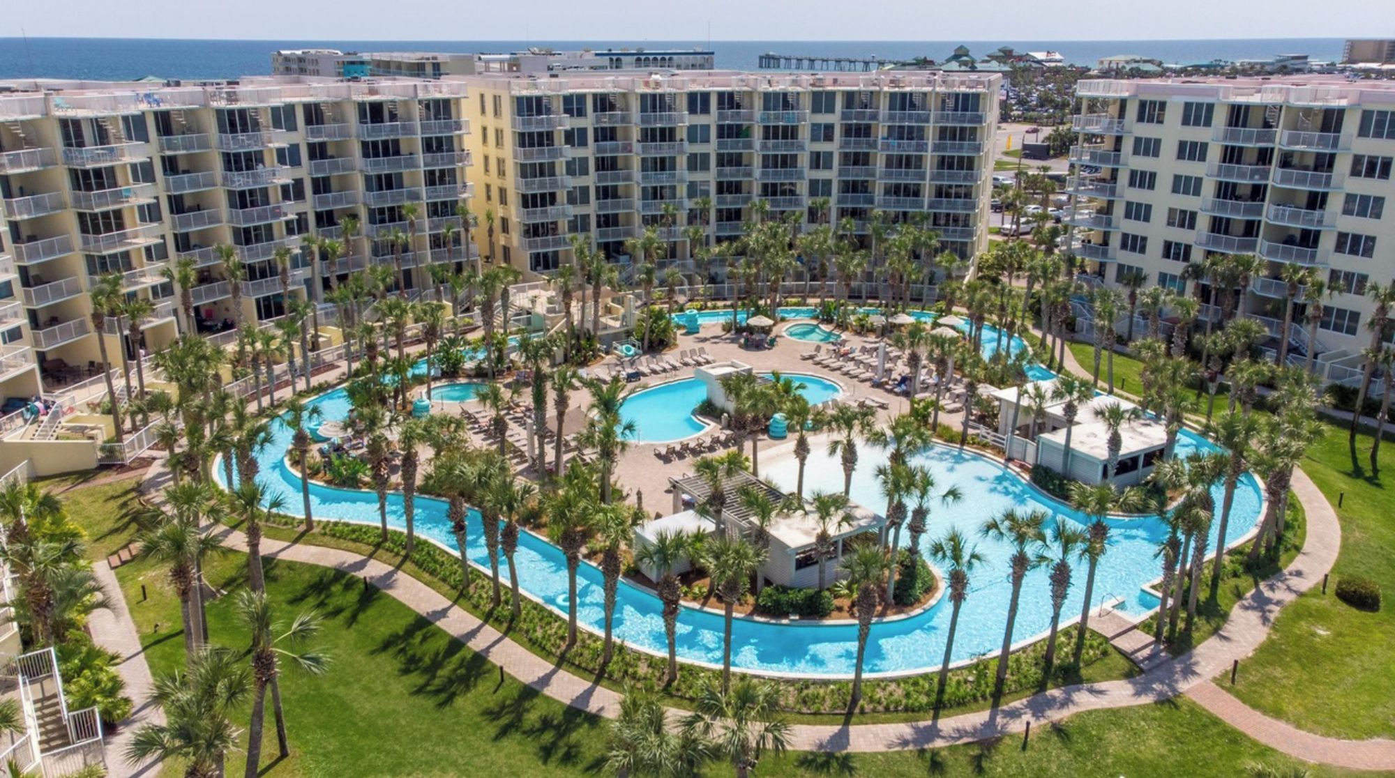 A hotel with multiple floors, a central pool area surrounded by palm trees, and a view of the ocean.