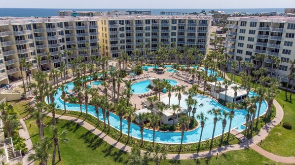 A hotel with multiple floors, a central pool area surrounded by palm trees, and a view of the ocean.