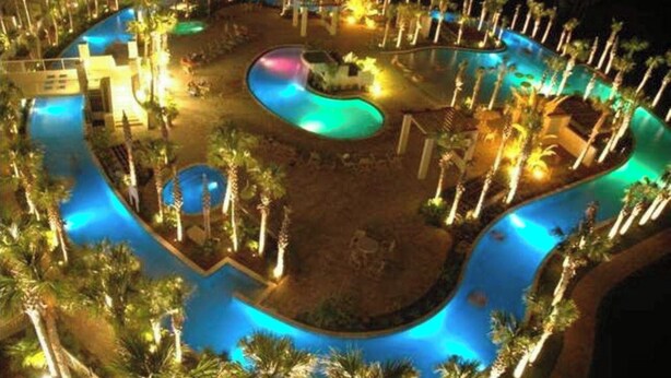 Een nachtzicht van een resort met een groot, kronkelend zwembad, omringd door palmbomen en verlicht met kunstlicht.