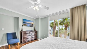 A bedroom with a large bed, a dresser, a chair, a ceiling fan, and a balcony with palm trees. - SANDPIPER 308-BAYSIDE CONDO @ DESTIN WEST SLEEPS 7 (Fort Walton Beach)