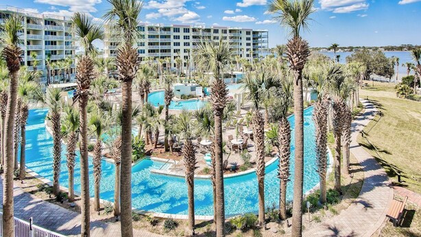 A resort with multiple swimming pools surrounded by palm trees, a large building in the background, and a clear blue sky.
