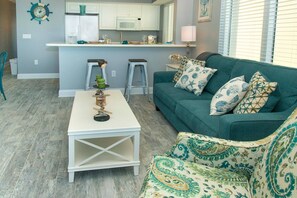 Ein Wohnzimmer mit einem türkisfarbenen Sofa, gemusterten Kissen, einem weißen Couchtisch und einer Küche im Hintergrund.