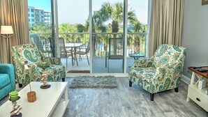 Un salon avec vue sur une piscine et des palmiers, comprenant des fauteuils à motifs, une table basse blanche et un canapé bleu.
