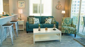 Un salon avec un canapé de couleur turquoise, un fauteuil à motifs, une table basse blanche et un petit coin bar avec des tabourets.