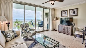 A spacious living room with a large sliding glass door leading to a balcony with a view of the ocean. There is a ceiling fan, a television, and a dining area with a table and chairs. - Sandpiper 508-1 BR+Bunkroom Corner Condo with Amazing views of Lazy River and Gulf! (Fort Walton Beach)
